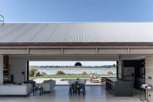 A spacious open-plan contemporary living, dining, and kitchen interior opening onto a waterfront view. The room features a large exposed timber roof structure, corrugated metal roof visible externally, extensive floor-to-ceiling glazing, pale timber flooring and wall lining, a dark kitchen island with bar seating, and a restrained neutral palette. A calm waterside landscape is visible beyond the deck.