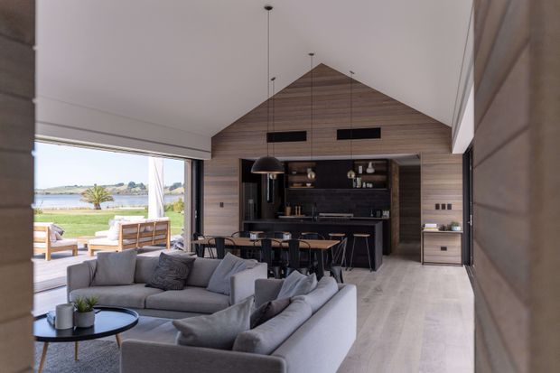 Open-plan contemporary living, dining and kitchen area with a dramatic vaulted timber-lined gable ceiling and wall, dark kitchen cabinetry, pendant lighting, pale flooring, and a large grey sectional sofa. Full-height glazed openings connect the room to a covered outdoor terrace and a rural landscape beyond.