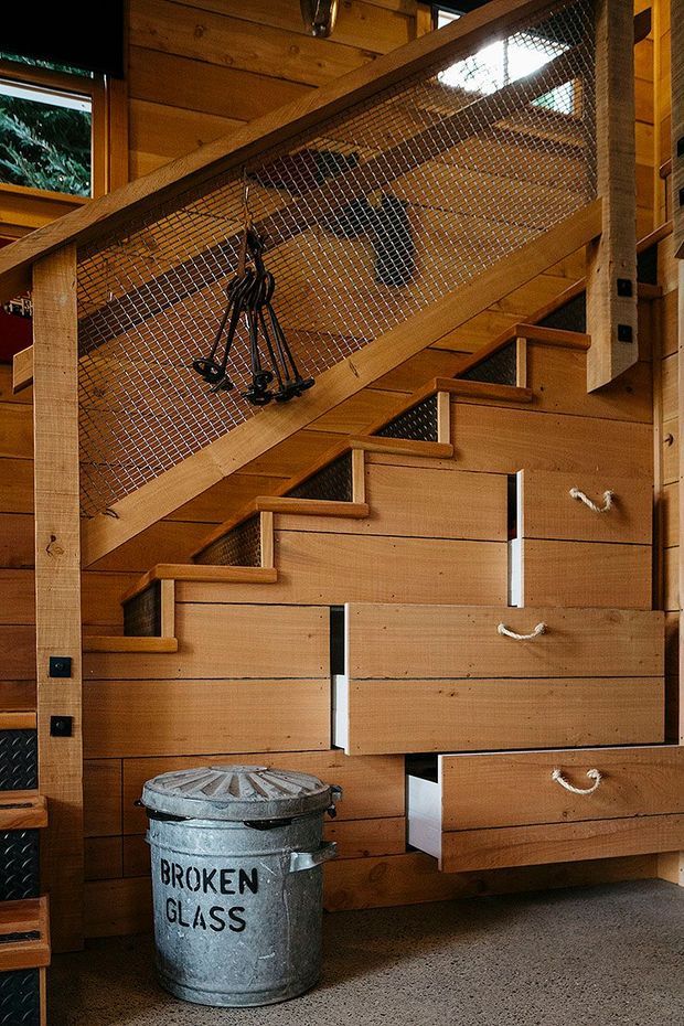 A compact timber-lined stairway with integrated pull-out drawers built beneath the steps. The staircase has warm wood treads and wall panelling, black metal mesh balustrading, and visible climbing or utility gear hanging from the railing. A metal recycling bin sits on the carpeted floor, emphasizing the practical storage-focused design.