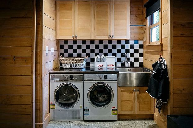 Compact laundry room with front-loading washer and dryer beneath light timber overhead cabinetry. The room features horizontal timber wall panelling, a black-and-white checkerboard splashback, a stainless-steel utility sink, and a small window with a dark roller blind.