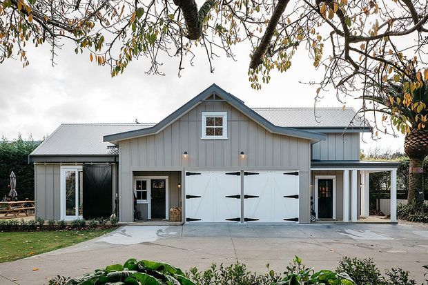 Front exterior view of a large grey weatherboard-style building with a prominent central gabled section, white corrugated pitched roofing, large white barn-style garage doors, and covered side verandas. The building is set behind a broad concrete driveway and landscaped garden, with mature trees framing the view.