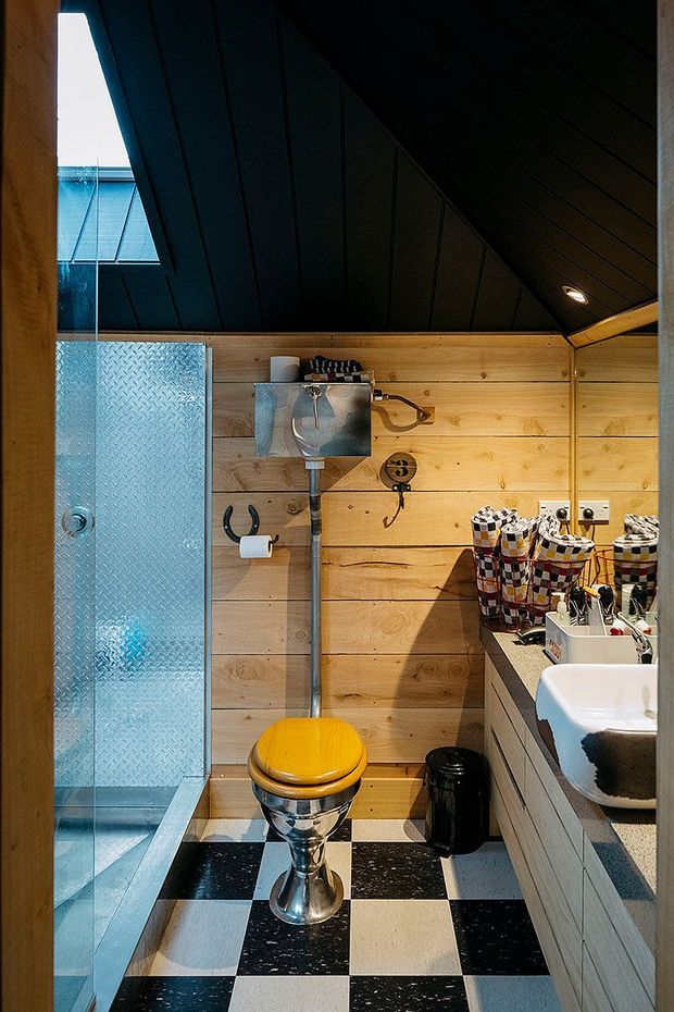 Compact eclectic bathroom with horizontal honey-toned timber wall panelling, a dramatic dark pitched ceiling, textured translucent glass shower enclosure, checkerboard black-and-white floor tiles, chrome plumbing, a pedestal toilet with bright yellow seat, and a white vanity with patterned storage containers. A roof skylight brings in natural light.