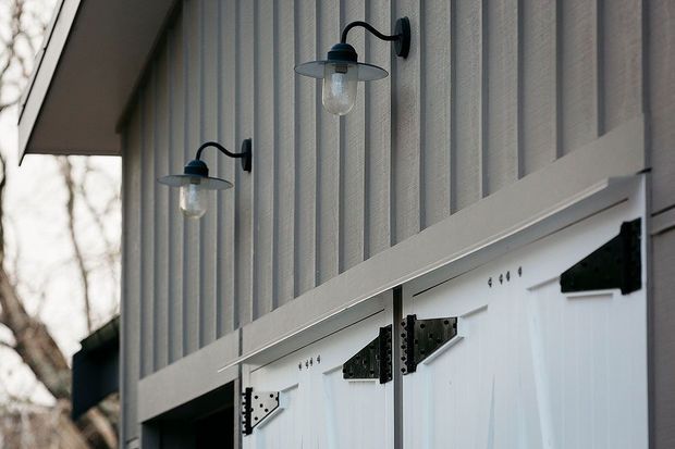 Close-up exterior view of a barn-style building facade with vertical grey weatherboard cladding, white framed barn doors, black strap hinges, and paired black wall-mounted gooseneck lights. The image emphasizes the rustic architectural detailing and material contrast.
