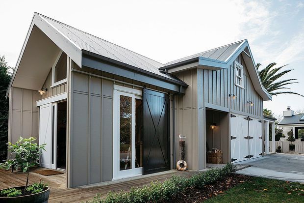 Exterior view of a contemporary barn-inspired home with grey vertical board-and-batten cladding, steep gabled metal roofs, tall glazed entry doors, and an integrated multi-car garage. The landscaped frontage includes a timber path, low garden beds, and lawn.