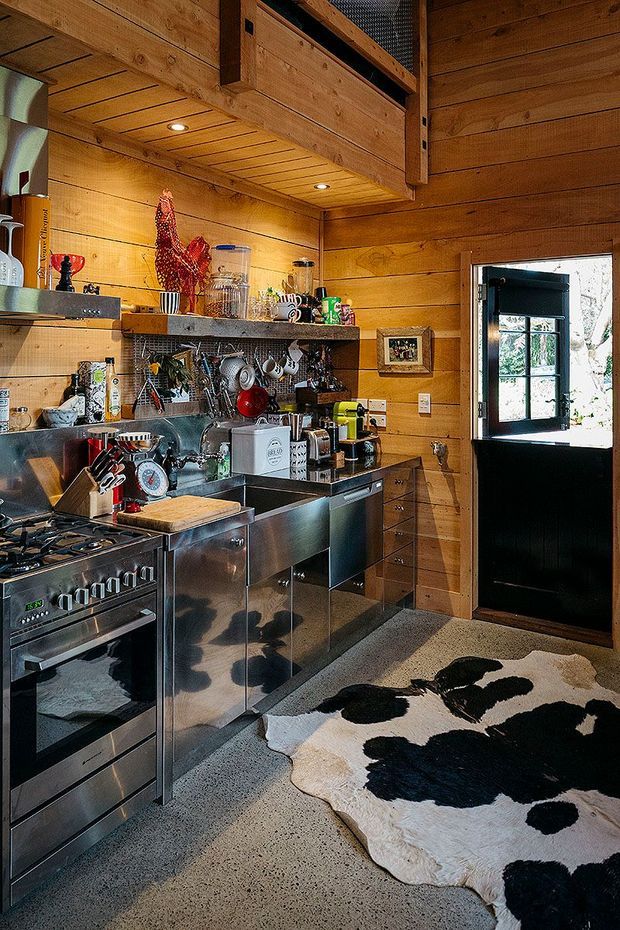 A compact rustic kitchen with extensive warm timber wall and ceiling panelling, exposed timber beams, stainless-steel cabinetry and appliances, open metal shelving, and a dark-framed glazed door. The space includes a gas range, sink, countertop appliances, utensils, and a cowhide rug over a speckled hard floor.