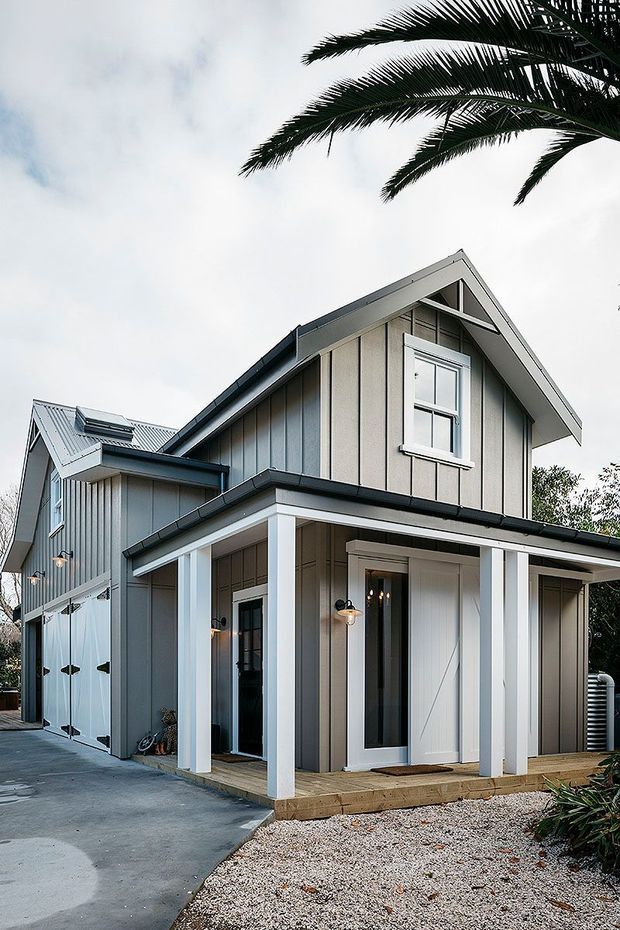Exterior view of a two-storey gabled home with a covered front porch, vertical light-grey weatherboard cladding, white trims and posts, dark metal roofing, and an attached garage. The home is set beside a paved driveway and gravel landscaping, with palm foliage visible overhead.