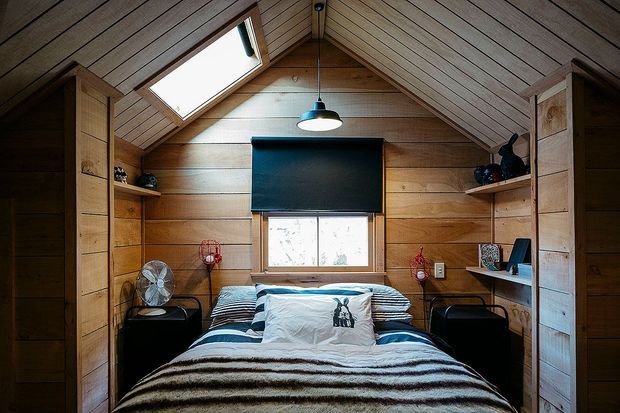 A compact attic-style bedroom with a peaked, timber-lined ceiling and matching horizontal timber wall panelling. The room contains a double bed with layered blue, white and brown bedding, built-in timber shelving and wardrobes on both sides, bedside tables, a central pendant light, and a skylight above. A roller blind covers the rear window, creating a warm, intimate cabin-like atmosphere.