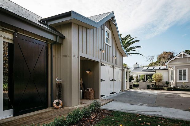 Exterior view of a coastal-style residential property with a weatherboard-clad, multi-gabled outbuilding or garage in the foreground. The structure features dark metal roofing, vertical board-and-batten wall cladding, large barn-style doors, and a covered connection toward a smaller cottage-like building. A paved driveway and landscaped garden sit between the buildings.