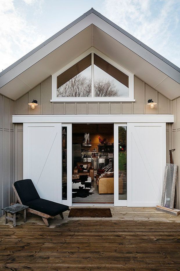 Exterior view of a compact gabled building opening onto a timber deck. The facade has light grey vertical board-and-batten cladding, white trim, a large triangular upper window, and wide white barn-style sliding doors. Through the open central doorway, a furnished living area is visible. A dark lounge chair and small outdoor table sit on the deck.
