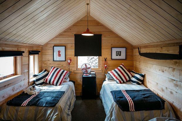 A compact timber-lined bedroom with a steeply pitched gable ceiling, two single beds, striped bedding and cushions, and a central wall-mounted television. The room features exposed warm wood panelling, a small central window, pendant lighting, framed artwork, and nautical or rustic decorative details.