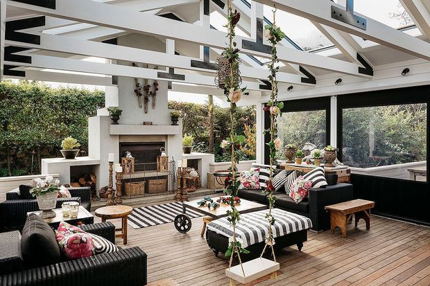 A bright, enclosed outdoor lounge or sunroom with a white painted timber-framed ceiling, exposed structural beams, and large black-framed glazed openings. The space features a central fireplace, striped black-and-white upholstered sofas and ottomans, timber decking, hanging plants, and a decorative hanging swing. It opens visually onto lush greenery and is furnished as a relaxed entertaining area.