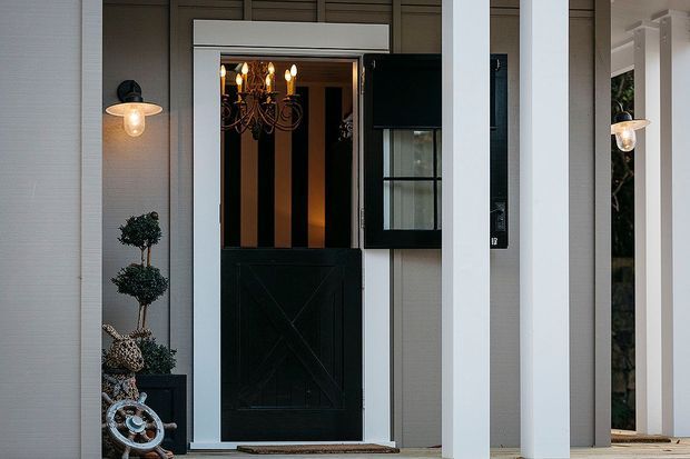 Close-up exterior view of a refined entryway with a black Dutch-style front door, glazed upper panels, and a visible chandelier inside. The entrance is framed by white vertical columns and dark grey wall cladding, with black wall sconces, a potted topiary, and nautical-style decorative hardware.