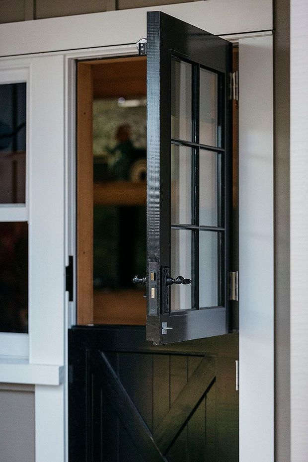 Close-up architectural detail of a partially open dark-painted timber entry door with divided glass panes, decorative black hardware, and a lower diagonal-braced panel. The door is set within white-painted exterior trim and weatherboard cladding, with a warm timber-lined interior visible beyond.