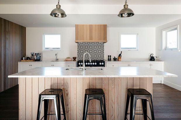 Bright contemporary kitchen with a large central island clad in vertical light timber boards and topped with a pale stone benchtop. Three black metal stools sit at the island, while white cabinetry lines the rear wall. A patterned dark tile splashback, timber rangehood surround, stainless pendant lights, and dark timber flooring add contrast.