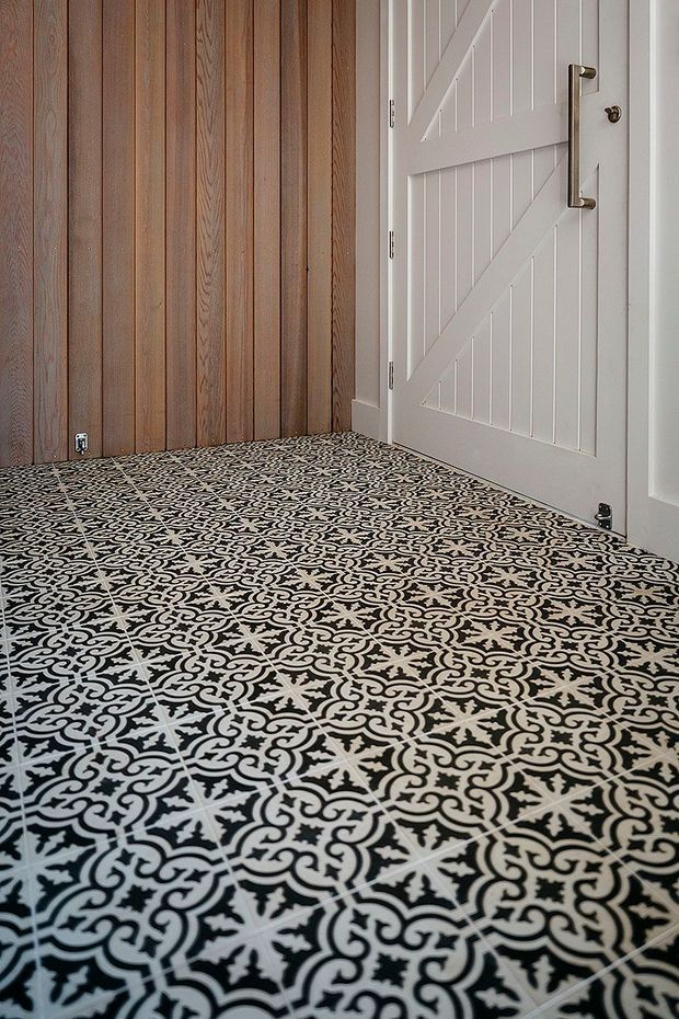 A small interior entry area featuring a highly patterned black-and-white tiled floor, vertical natural timber wall panelling, and a white barn-style door with diagonal bracing and brass hardware.