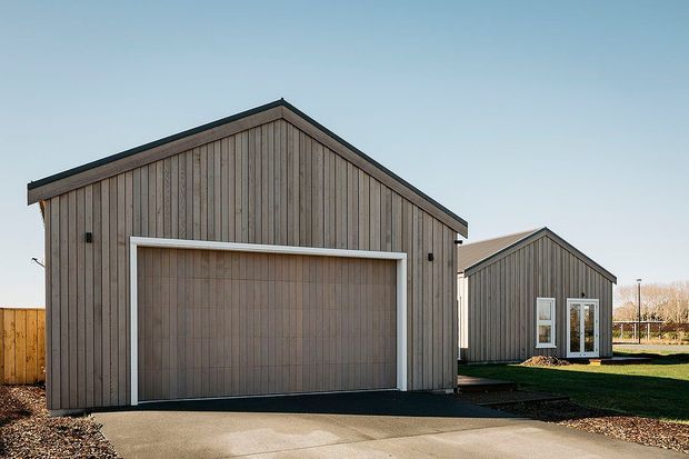 Exterior view of two simple gable-roofed buildings clad in vertical weatherboard timber, with a large garage door on the foreground structure and glazed doors/windows on the smaller rear structure. The buildings sit beside a paved driveway and lawn under a clear sky.