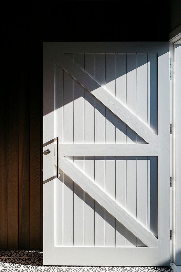 Close-up architectural detail of a white vertically paneled timber door with prominent diagonal and horizontal bracing, a simple metal handle, and visible hinges. The door is set within a dark timber-lined opening and opens onto patterned tile flooring. Strong sunlight creates geometric shadows across the door surface.