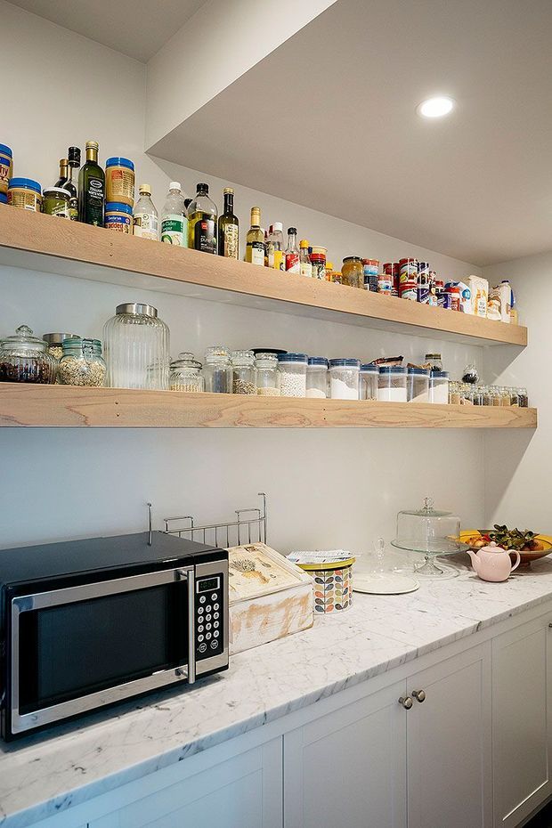 A compact, well-organised pantry or kitchen storage area with two long open timber shelves displaying bottles, jars, tins and pantry goods. Below, white cabinetry supports a white marble-look benchtop with a microwave, dishware, a cake stand and fruit bowl. The space has white walls, recessed ceiling lighting and a bright, practical fit-out.