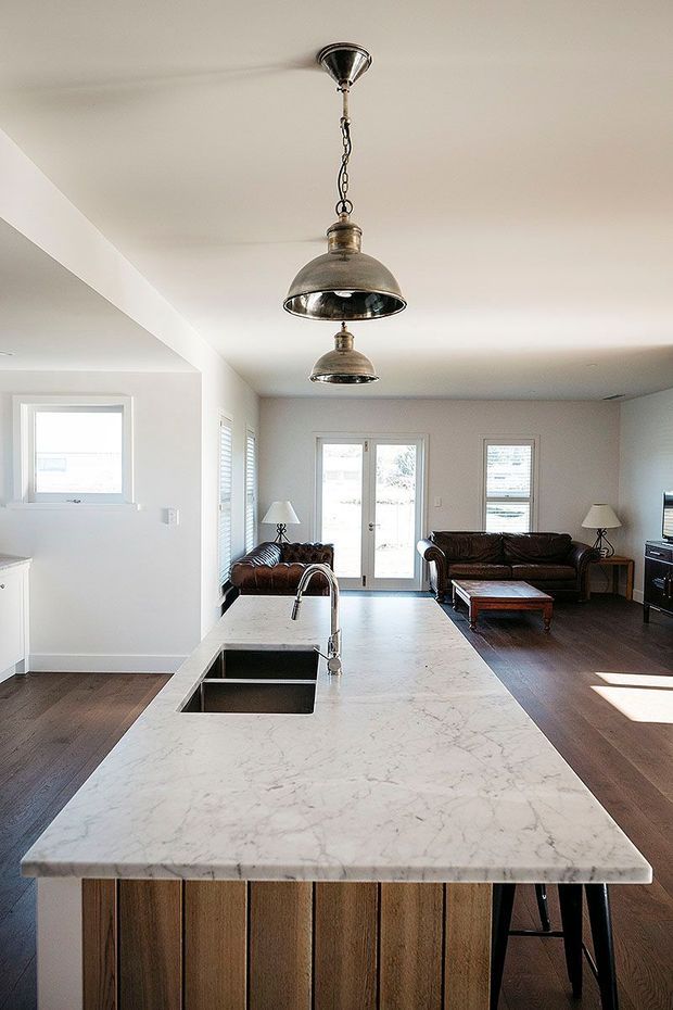 Open-plan interior viewed from a large kitchen island, with a white marble-look benchtop, double undermount sink and curved tap. The island has vertical timber panelling and seating. A bright living area with dark timber flooring, leather sofas, lamps, shutters and pendant lights is visible beyond.