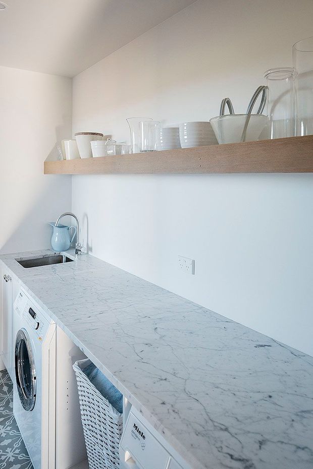 Compact, bright laundry room with a long white marble-look benchtop, integrated front-loading appliances, a deep sink with curved faucet, pale timber open shelving, and white ceramic and glassware displayed above. Patterned floor tiles add a subtle decorative detail.