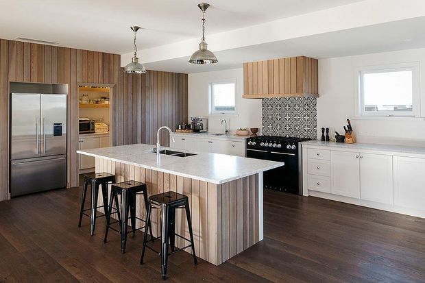 Bright contemporary kitchen with a large white island and integrated sink, four black metal bar stools, white cabinetry, stainless-steel appliances, vertical timber wall panelling, patterned tile splashback, dark timber flooring, and three industrial-style pendant lights.