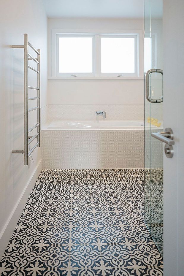 Bright, narrow contemporary bathroom with a white bathtub beneath two frosted or overexposed windows, a chrome heated towel rail, glass shower screen and door, white wall surfaces, and highly patterned black, white, and grey floor tiles. The room has a clean, high-contrast decorative finish.