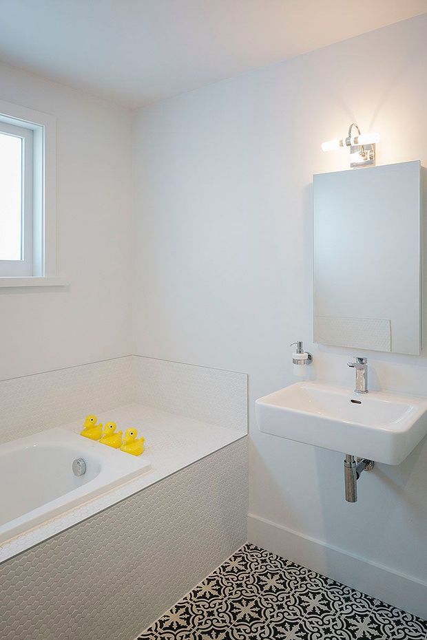 Bright, compact bathroom featuring a white bathtub with a tiled surround, a wall-mounted white basin, mirrored cabinet, chrome tapware, patterned black-and-white floor tiles, and a small window. Yellow rubber ducks add a playful accent.