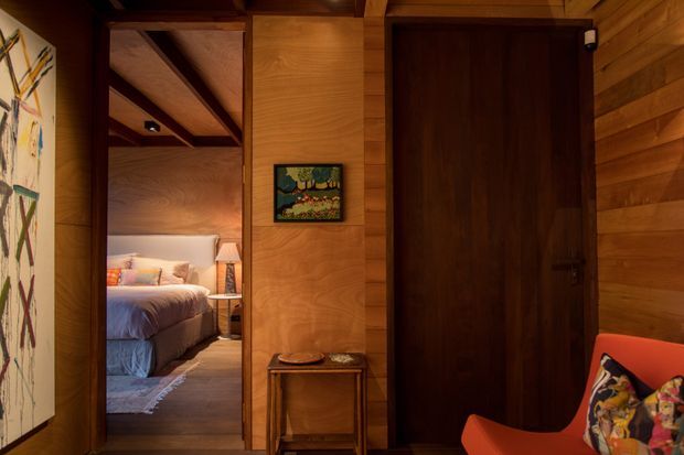 Warm, moody interior view dominated by extensive timber wall panelling, exposed timber ceiling beams, and a dark timber door. A bedroom is visible through an open doorway, with a small console table and artwork in the foreground and part of a bright orange lounge chair at right.