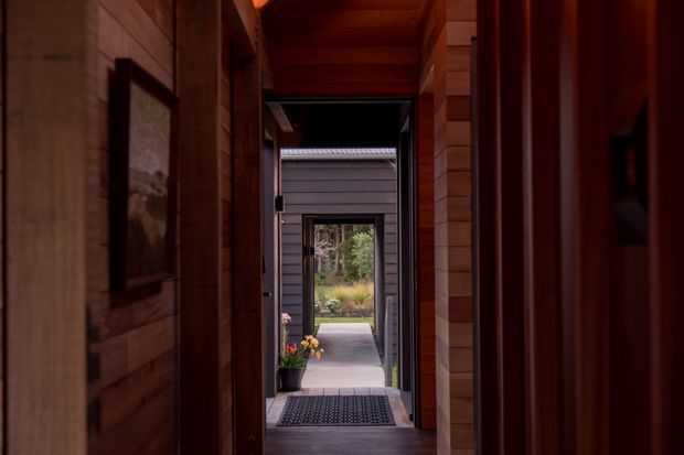 A narrow, warmly lit timber-lined hallway or entry passage leads toward an open exterior doorway. The corridor has rich dark wood wall and ceiling cladding, a framed artwork on the left, and a dark entrance mat near the threshold. Through the aligned doorways, a garden and path are visible.