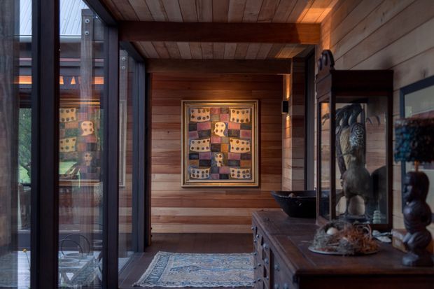 A warm, dimly lit timber-lined interior hallway or entry passage. Horizontal timber wall boards and a timber-planked ceiling are framed by substantial dark exposed beams. Full-height glazing and dark framed doors line the left side, while a dark timber console or built-in display cabinet with sculptural objects occupies the right. A patterned framed artwork hangs at the end wall, and a traditional rug defines the floor. The space has a refined, gallery-like character with strong natural-material detailing.