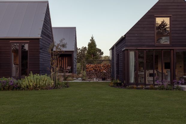 A small group of dark timber-clad gabled buildings arranged around a landscaped lawn and central firewood stack. The buildings feature pitched metal roofs, large glazed doors and windows, and a restrained cabin-like architectural character in a wooded setting.