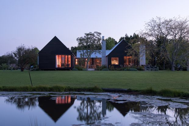 A contemporary dark-clad residence set within expansive landscaped grounds beside a reflective pond. The building comprises several steeply pitched gabled volumes with warm interior lighting, large glazed openings, and prominent pale chimney-like elements. The dusk setting creates strong reflections in the water and a calm, atmospheric composition.