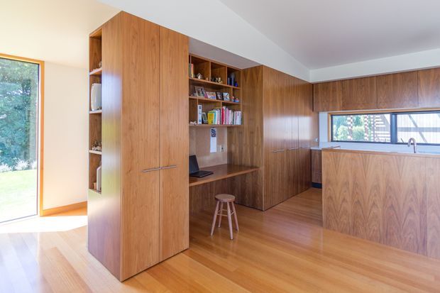 A warm, timber-lined open-plan interior combining a compact kitchen with integrated cabinetry, shelving, and a built-in study nook. Floor-to-ceiling walnut-toned joinery forms a central storage wall and continues around the kitchen, with a matching island featuring a raised benchtop and undermount sink. Honey-toned timber flooring, white ceilings, and abundant glazing create a bright, cohesive space.