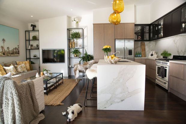 Bright open-plan kitchen and living area with dark timber flooring, white walls, warm wood cabinetry, a large marble-look island, black-framed glazing and shelving, stainless steel appliances, and amber pendant lights. A living room with sofa, TV, shelving and coffee table is visible beside the kitchen, creating a warm contemporary family space.