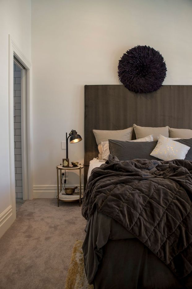 A cozy, softly lit bedroom with a large dark upholstered or wood-toned headboard, layered charcoal and taupe bedding, neutral walls, and carpeted flooring. A small metal bedside table holds a warm-glowing lamp and accessories. A bathroom or tiled wet-area doorway is visible at the left, while a dark textured round wall decoration provides a bold focal point above the bed.