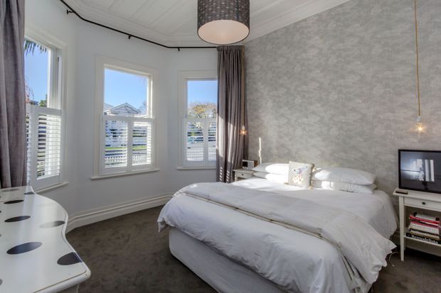Bright, elegant bedroom with a large upholstered bed dressed in white linens, grey carpet, textured grey feature wall, white-painted trim and ceiling, tall bay-style windows with plantation shutters, and full-length grey curtains. A curved white built-in desk or console occupies the foreground, while a small shelving unit and television sit beside the bed. The room has a refined, contemporary-traditional feel with abundant natural light.