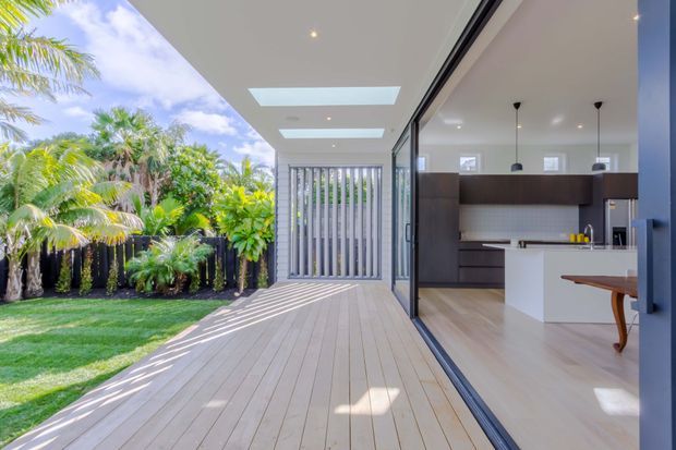Bright contemporary indoor-outdoor living space with a pale timber deck opening directly into a modern kitchen through fully glazed sliding doors. The kitchen features dark wood cabinetry, a white island, pale flooring, pendant lights and a tiled splashback. A lush tropical garden and slatted privacy screen are visible beyond the deck, while skylights and recessed lighting bring additional daylight indoors.