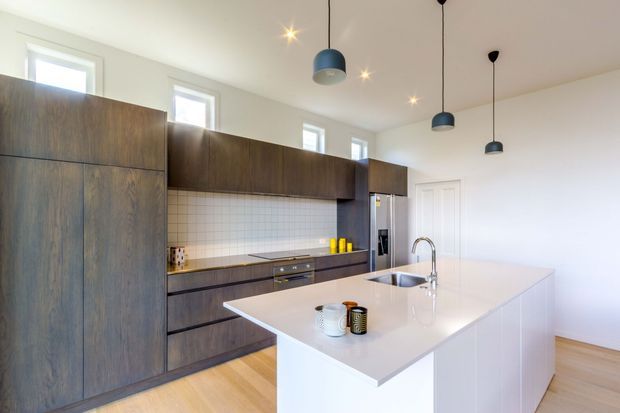 Bright contemporary kitchen with a large white island, dark timber-look full-height cabinetry, pale square-tile splashback, integrated appliances, light timber flooring, and multiple black pendant lights. High-level windows bring natural light into the room.