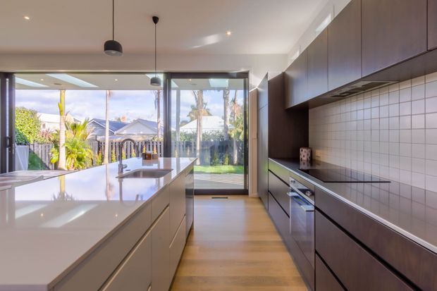 A bright contemporary galley kitchen with a large central island, white stone benchtops, dark wood cabinetry, integrated appliances, and a white square-tile splashback. Full-height glazing and a glass door open to a leafy outdoor area, bringing in abundant natural light.