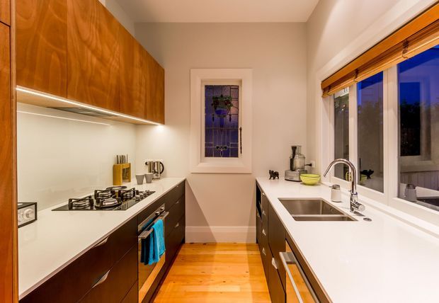 A compact galley kitchen with warm timber cabinetry, white benchtops and backsplash, integrated under-cabinet lighting, a gas cooktop and oven on the left, and a double-bowl stainless-steel sink beneath a large window on the right. The room has polished timber flooring, a small stained-glass window, and a warm, refined interior.