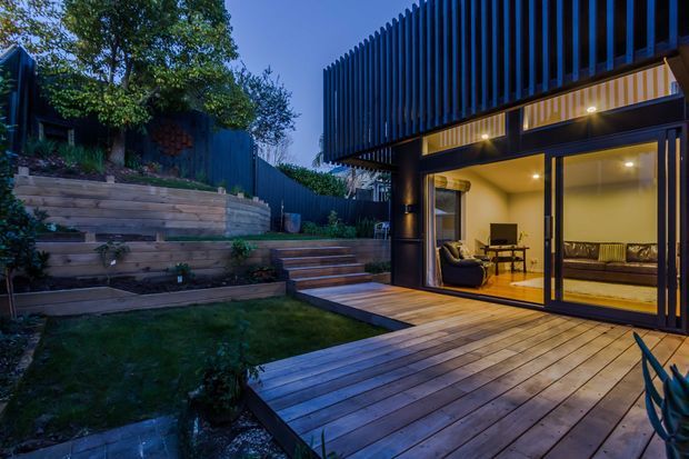 Twilight exterior view of a modern multi-level home addition opening onto a timber deck and landscaped terraced garden. The dark facade features strong vertical screening, broad glazed sliding doors, warm interior lighting, and a cantilevered upper level. The visible interior appears to be a living room with seating and a dining table.