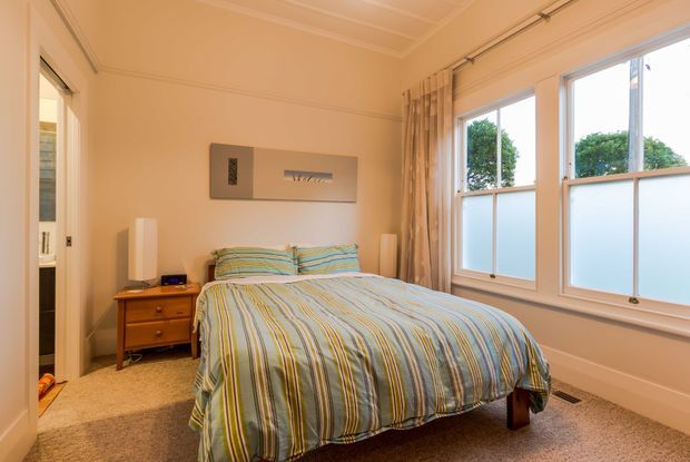 A warm, traditional bedroom with a neatly made bed dressed in striped blue, green, and cream bedding. The room features beige walls, carpeted flooring, a timber bedside table, framed wall art, sheer curtains, and large windows with frosted lower panes. A doorway and built-in storage are visible at the left.