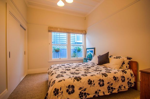 A compact, warmly lit bedroom with cream walls, decorative ceiling and wall mouldings, a carpeted floor, built-in sliding wardrobe, and a double bed dressed in a floral-patterned duvet with layered pillows. A shuttered window provides natural light, with a small bedside cabinet and framed artwork adding detail.