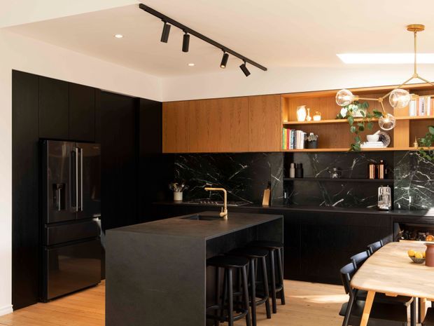 A sophisticated contemporary kitchen with a large dark stone island, black cabinetry and appliances, rich timber overhead cabinetry and open shelving, and a dramatic black marble-look splashback. The island includes seating for several people, while a timber dining table sits adjacent to the kitchen. Track lighting and globe pendant lights provide layered illumination.