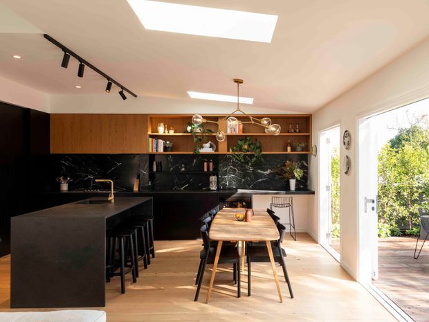 A bright open-plan kitchen and dining area with dark, dramatic cabinetry and a large black marble-look island and splashback. Open timber shelving displays books and decor, while a timber dining table sits beneath a sculptural globe pendant. Skylights and full-height glazed doors bring in abundant natural light and connect the room to an outdoor deck.