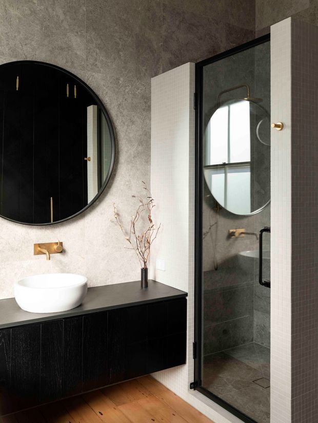 Contemporary bathroom featuring a wall-mounted black timber vanity with a white vessel basin, brass wall-mounted tapware, a large round black-framed mirror, and a frameless black-framed glass shower enclosure. Textured grey stone-look wall surfaces contrast with small white mosaic tile detailing, while warm timber flooring adds natural warmth. The shower includes a round mirror and brass fixtures visible through the glass.