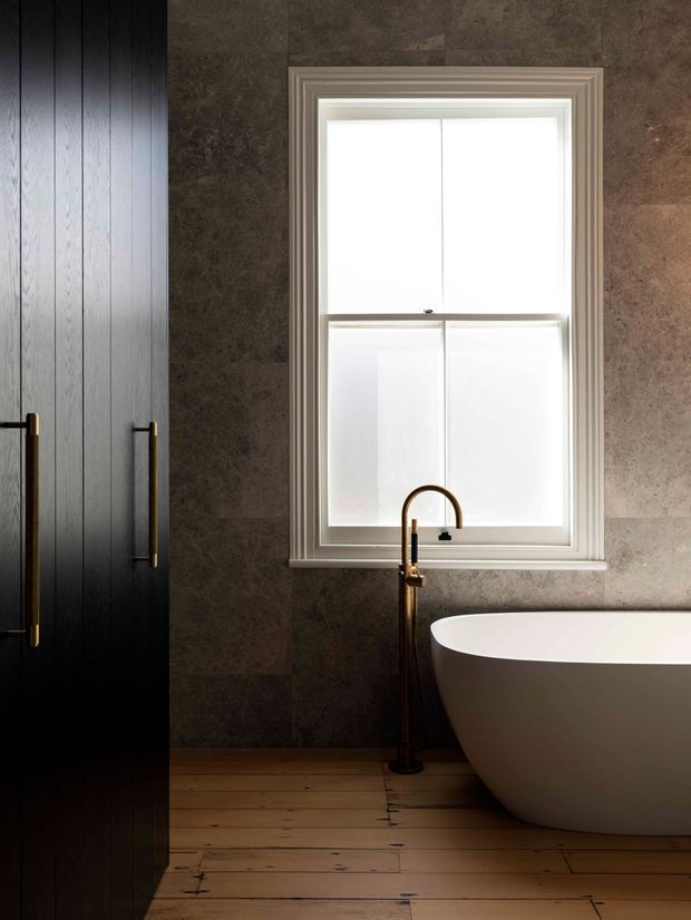 A moody, refined bathroom featuring a freestanding white bathtub beneath a large frosted-glass window, a tall dark vanity or storage unit with slim brass handles, a brass-toned floor-mounted bath tap, dark stone-look wall tiling, and warm timber floorboards.
