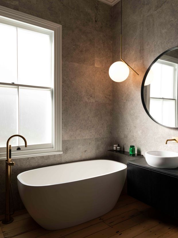 A refined bathroom featuring a freestanding white oval bathtub beneath a large frosted window, a dark timber vanity with a white vessel basin, a round black-framed mirror, and a globe pendant light. The walls are clad in textured grey stone-look panels, while warm timber floorboards and brass-toned tapware add contrast.