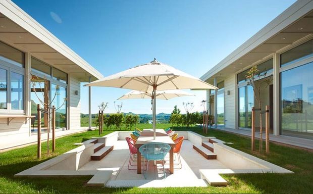 A bright landscaped courtyard between two low, modern pavilion-like wings. The central paved outdoor dining area has a large cream umbrella, glass-top table, colourful chairs, stepped built-in seating, and white rendered or concrete planter walls. Floor-to-ceiling glazing and pale horizontal cladding define both adjoining buildings, with a rural landscape visible beyond.