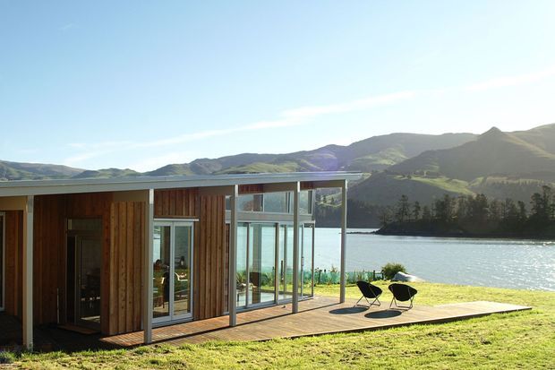 A single-storey modern lakeside dwelling with a low flat roof, extensive floor-to-ceiling glazing, and warm vertical timber cladding. A timber deck with two black lounge chairs extends toward the water, with mountainous terrain and trees in the background.